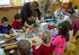 Dzieci przy ozdobionym świątecznie stole rozwałkowują ciasto na pierniki oraz przy użyciu foremek formują pierniki.